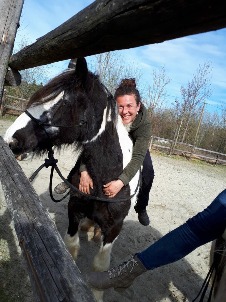 Eine glücklich lächelnde Frau sitzt auf einem schwarzen-weißen Pferd und umarmt seinen Hals.