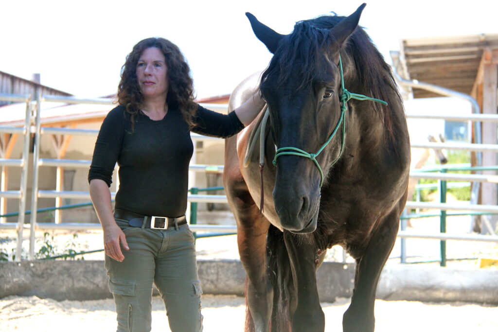 Eine Frau steht neben einem schwarzen Pferd und hat ihre Hand auf seinen Widerrist gelegt.