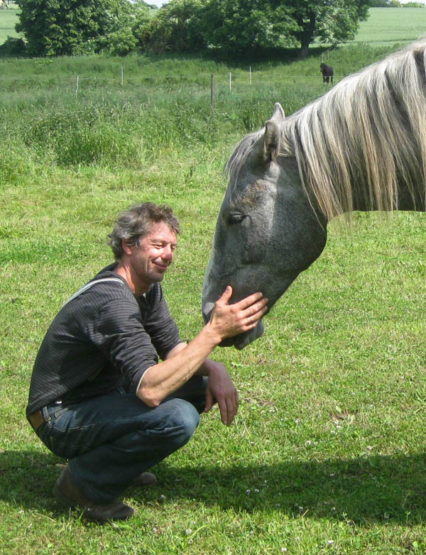 Ein Mann sitzt in der Hocke lächelnd vor einem großen grauen Pferd, das sein Gesicht in die offene Hand des Mannes schmiegt.