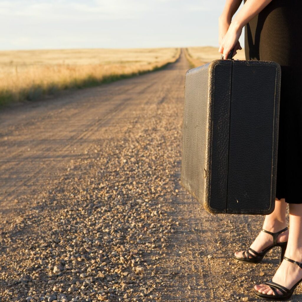 Eine Frau mit einem Koffer in einer kargen Landschaft, die von einer Straße durchschnitten wird.