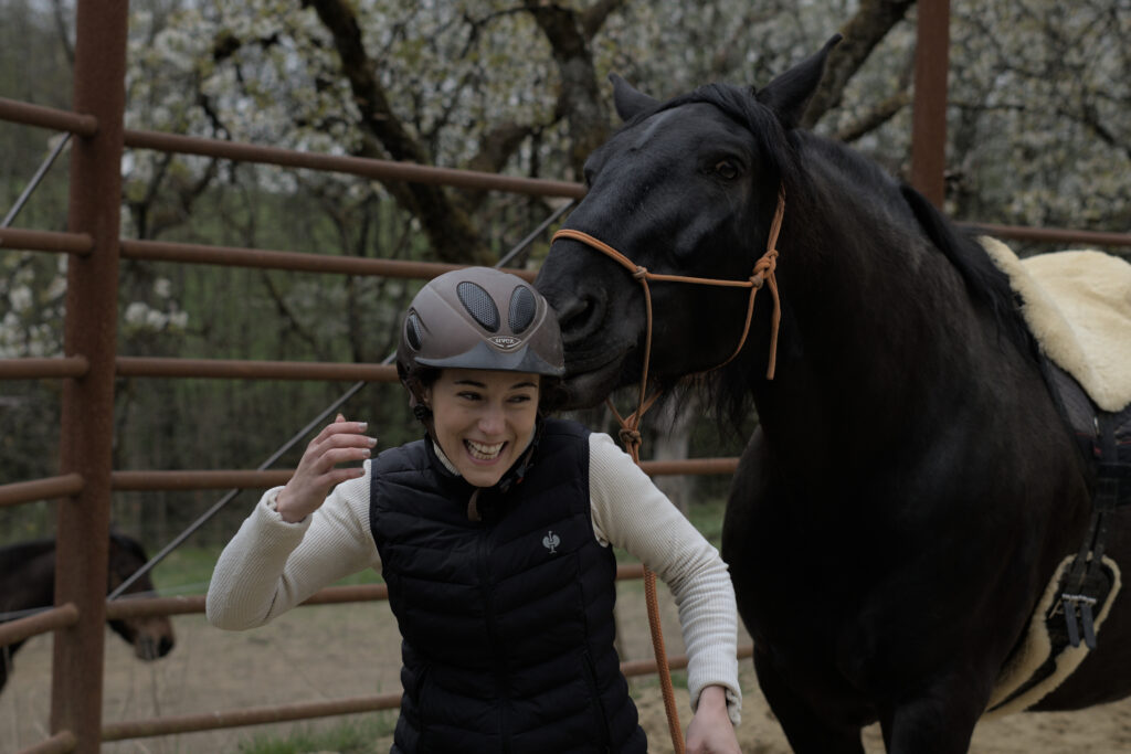 Eine junge Frau, die ein schwarzes Pferd führt, das versucht, mit ihrem Reithelm zu spielen. Die Frau lacht, ist aber beunruhigt.