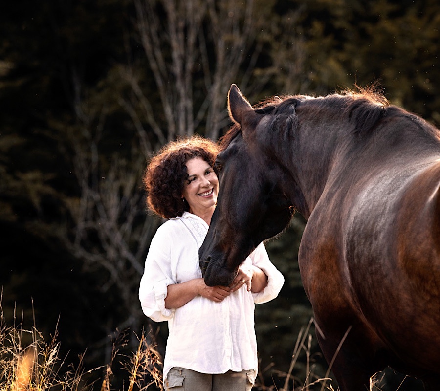Ein großes schwarzes Pferd legt seine Schnauze auf die Hände einer Frau mit weißer Bluse, die ihn anlächelt.