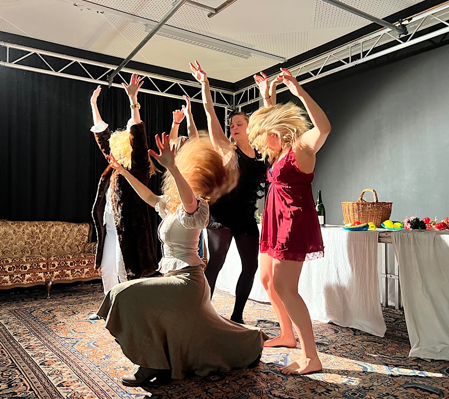 Wild tanzende Frauengruppe in einem Bühnenbild mit Sofa und weiß gedecktem Tisch.