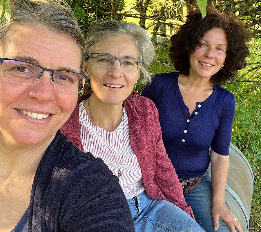 Drei Frauen sitzen hintereinander auf einem Metallfass und lächeln in die Kamera.