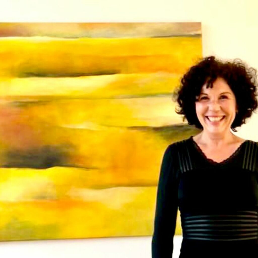 Eine schwarz gekleidete Frau lächelt breit vor einem gelben abstrakten Gemälde.