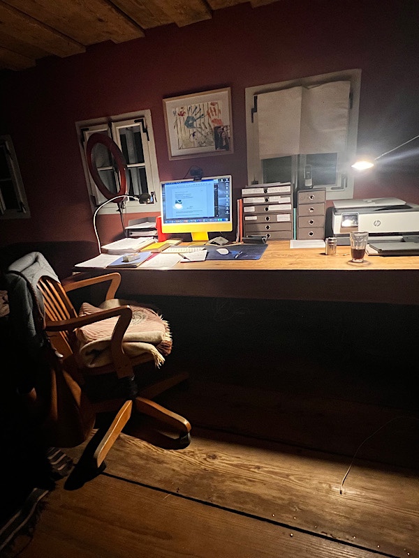 Ein dunkler Arbeitsraum, in dem nur Tisch und Arbeitsstuhl beleuchtet sind. Auf dem Tisch ein Computer und Papiere, Drucker, Ablagen.