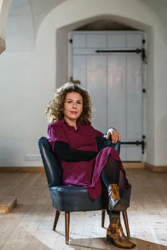 Elma Esrig auf einem Sessel im Hintergrund eine Tür. Elma hat ein lilafarbenes Kleid an und schlägt die Beine übereinander.