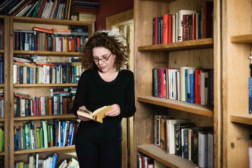 Elma Esrig neben und vor einer Bücherwand. Sie ist schwarz gekleidet, trägt eine Brille und blättert stehend in einem alten Buch.