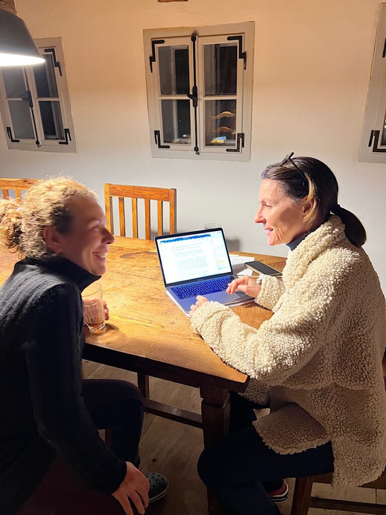 Zwei Frauen an einem großen Holztisch, auf dem ein Laptop steht, unterhalten sich lächelnd.