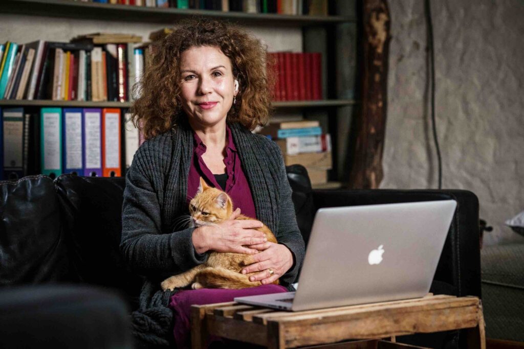 Elma Esrig, in einem lila Kleid und Strickjacke, sitzt vor ihrem Laptop mit Kopfhörern im Ohr und hält eine rotgetigerte Katze im Arm.