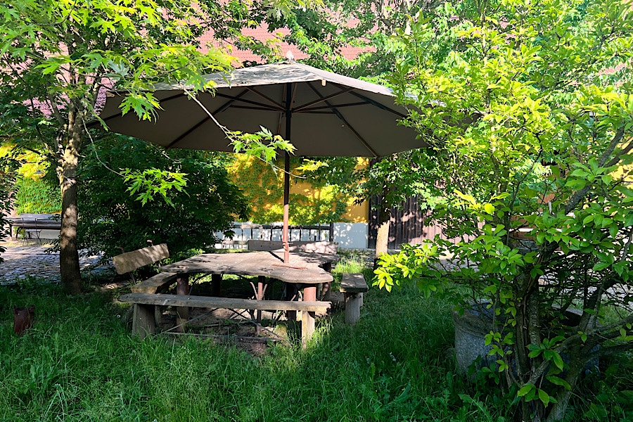 Ein Schattenplatz im Innenhof mit kleiner Wiese, großem Tisch und Bänken und einem großen Sonnenschirm.