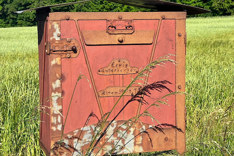 Ein rötlich angestrichener selbstgebauter Briefkasten aus Holz und Eisen vor einer hohen Wiese.