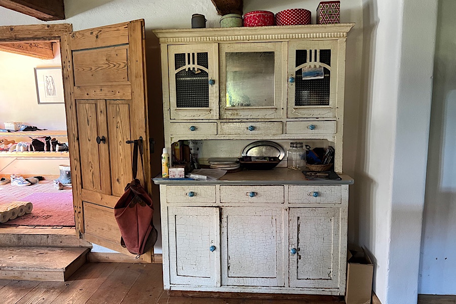 Ein altmodischer weißer Vorratsschrank in einer Küchenecke, daneben eine zum Flur hin geöffnete Tür.